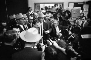 Lee Harvey Oswald is surrounded by reporters at a midnight press conference held in the basement assembly room of Dallas police headquarters.